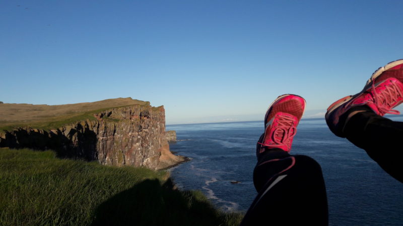 W kadrze znajdują się ludzkie stopy w czerwonych sportowych butach do biegania uniesione na tle nadmorskiego pejzażu z wyraźnie nakreśloną linią horyzontu